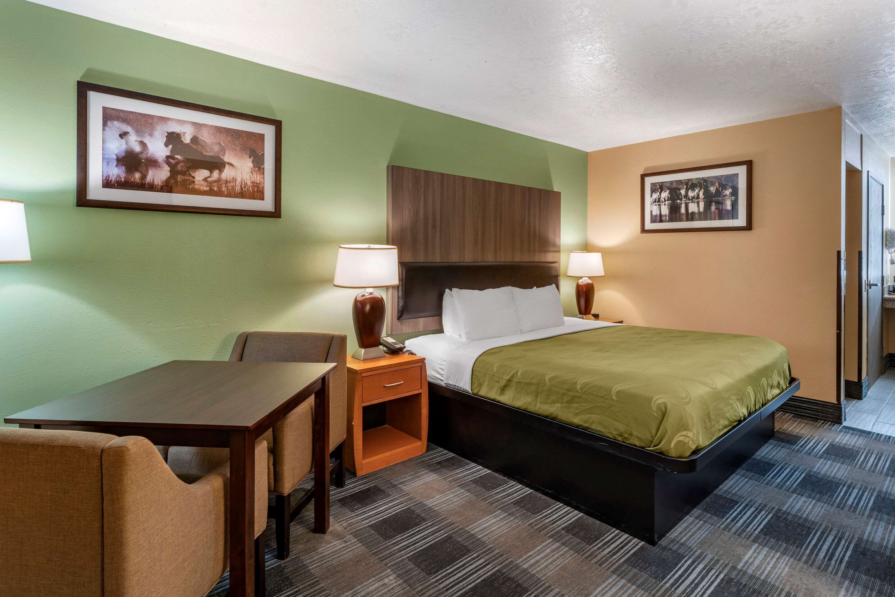 Habitación de hotel con cama, escritorio, dos sillas, mesita de noche con lámparas y dos cuadros en las paredes.