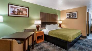 Habitación de hotel con cama, escritorio, dos sillas, mesita de noche con lámparas y dos cuadros en las paredes.