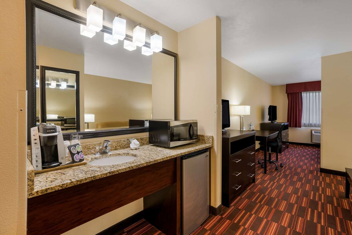 Quarto de hotel com cozinha compacta, um espelho grande, cafeteira, micro-ondas, frigorífico, secretária com cadeira e janela com cortinas.