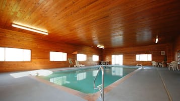 An indoor swimming pool with wooden walls and ceiling, surrounded by lounge chairs and tables.