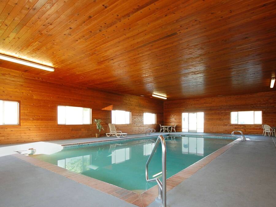 An indoor swimming pool with wooden walls and ceiling, surrounded by lounge chairs and tables.