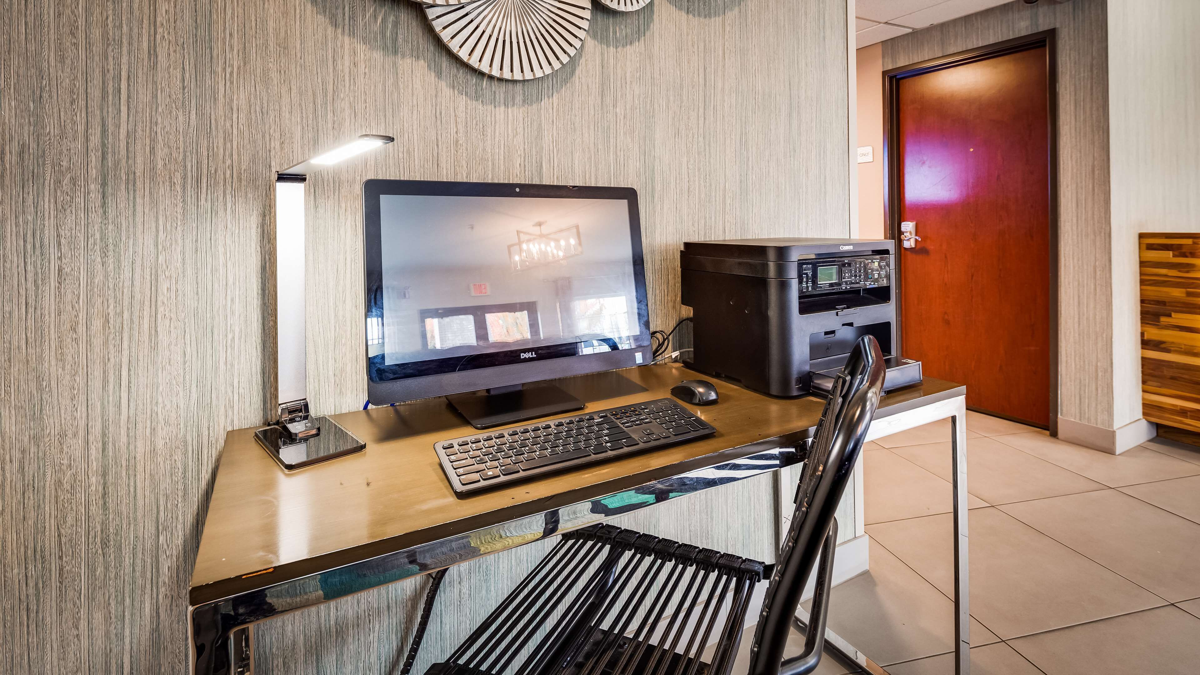 Un escritorio moderno de oficina con un monitor Dell, teclado e impresora, frente a una pared de madera con una puerta al fondo.