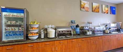 Espaço de café da manhã de hotel com geladeira repleta de bebidas e frutas, buffet variado e estação de café.