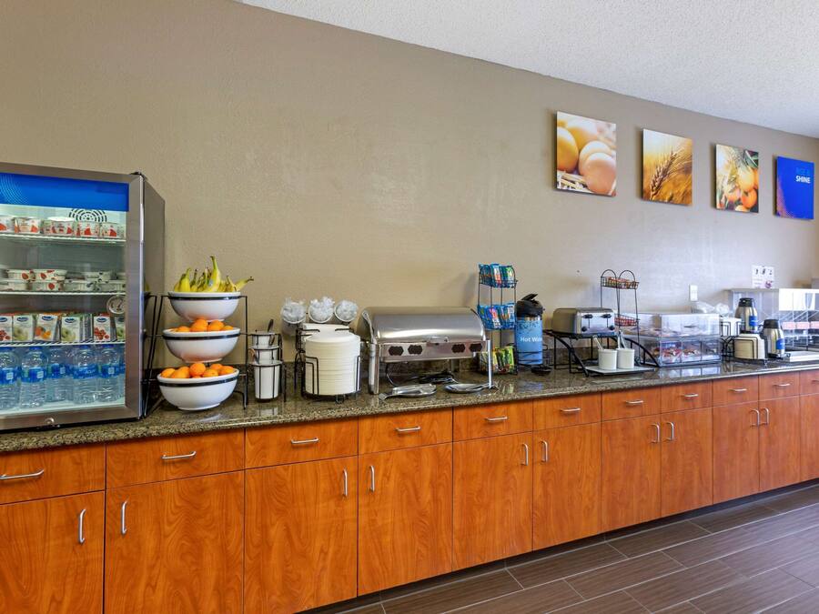 A hotel breakfast area with a refrigerator stocked with beverages and fruits, a buffet setup with various food items, and a coffee station.