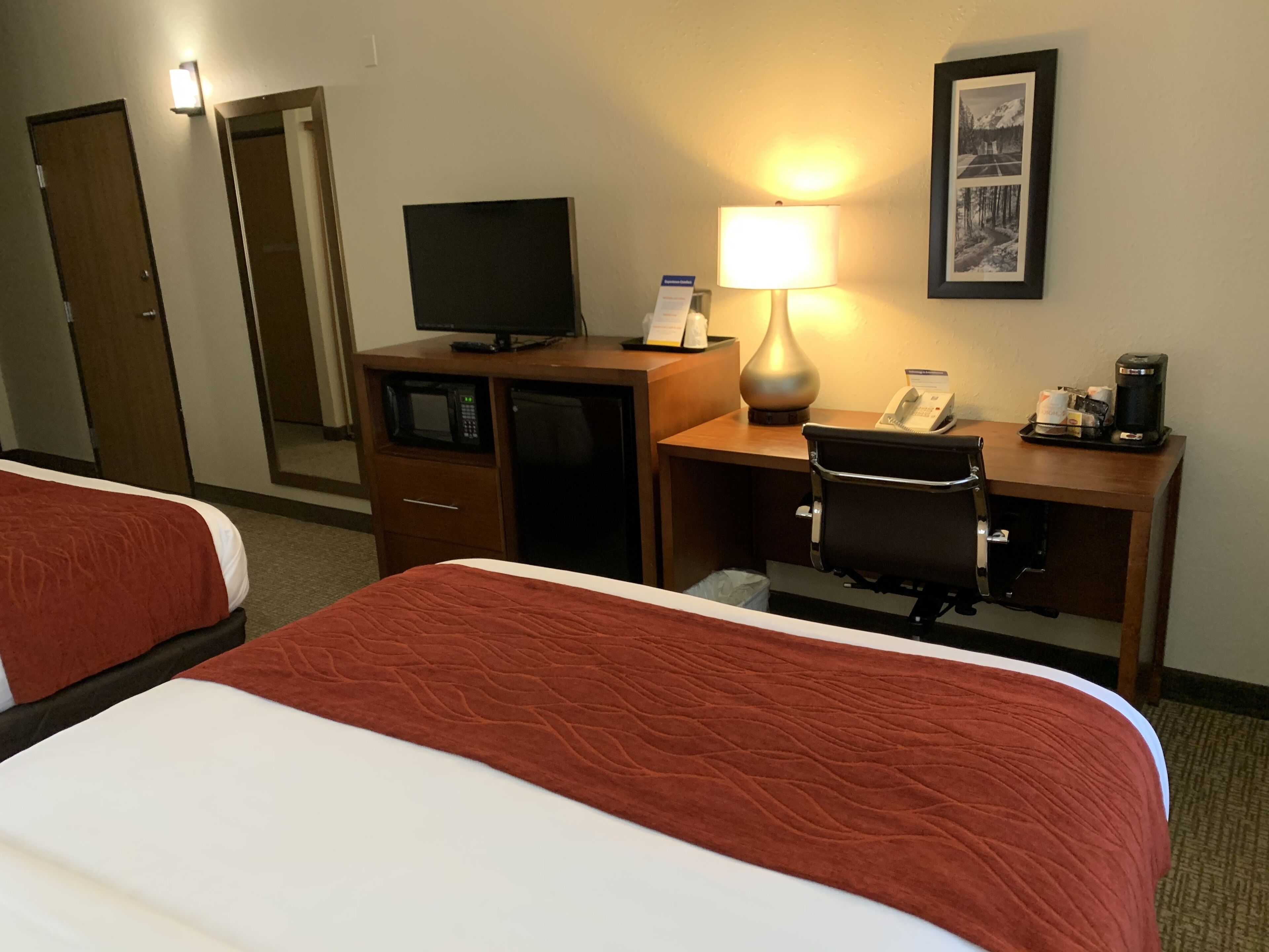 A hotel room with two beds, a desk with a computer, a lamp, a chair, a coffee maker, and a framed picture on the wall.