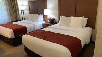 A hotel room with two beds, each with a wooden headboard, white bedding, and a red accent. There are bedside tables with lamps and a small potted plant.