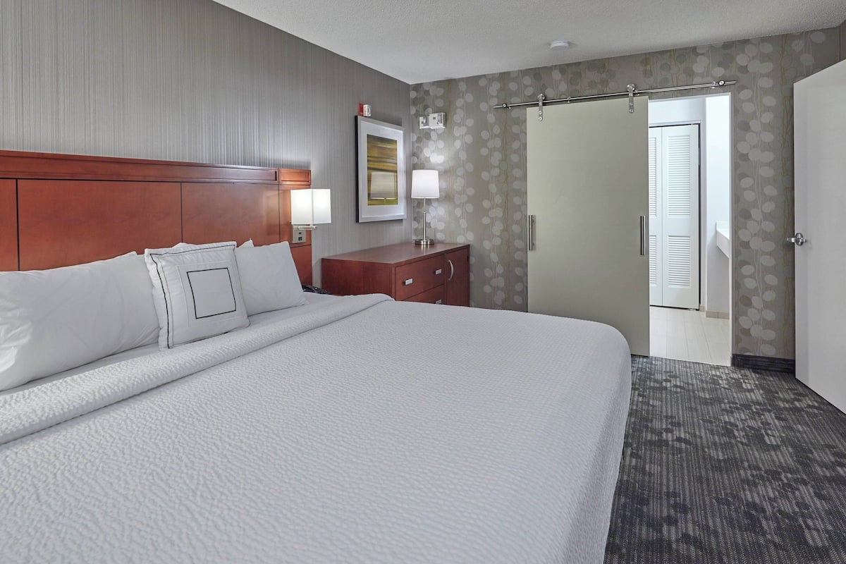 Habitación de hotel con una cama grande, cabecera de madera, mesita de noche con lámpara, cómoda y puerta que da a un pasillo.