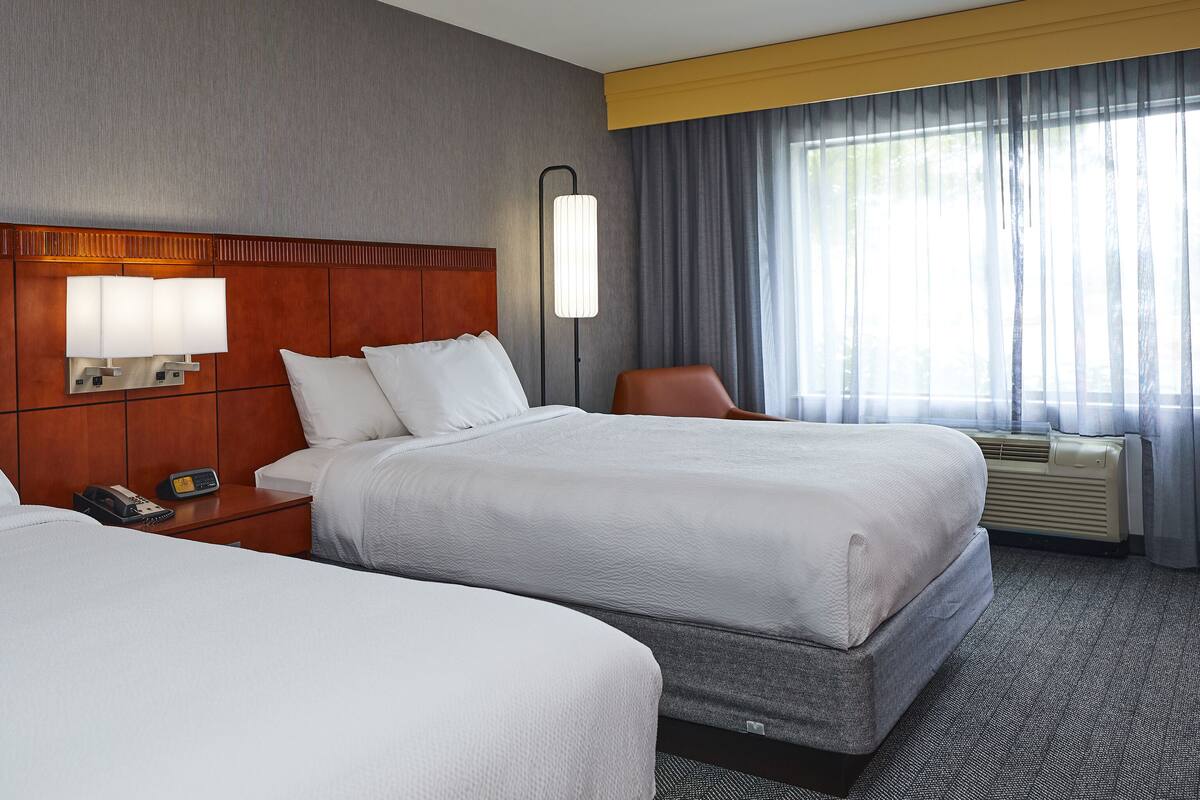 Habitación de hotel con dos camas, una mesita de noche, una lámpara, una silla y un ventanal con cortinas.