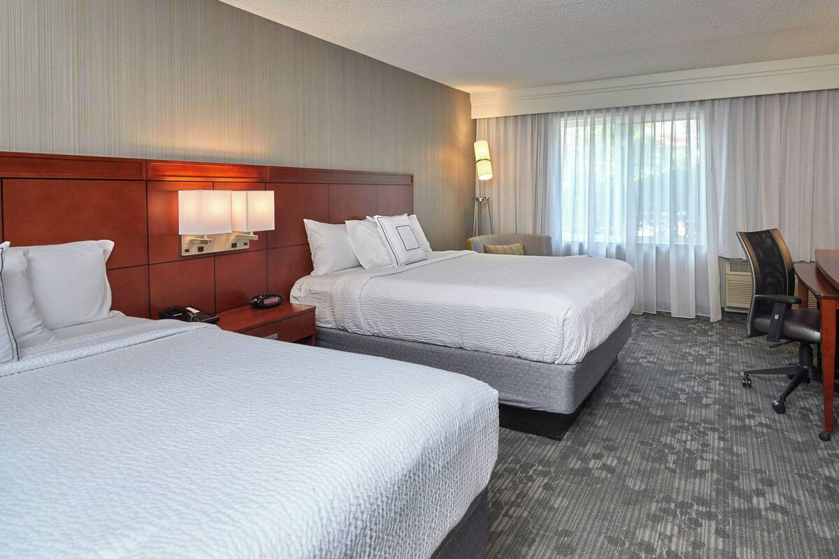 Habitación de hotel con dos camas, un escritorio, una silla, una lámpara y una ventana con cortinas.
