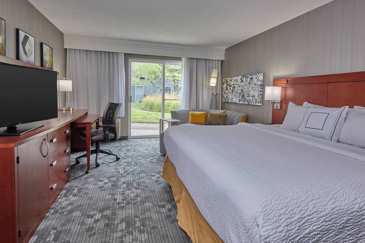 Habitación de hotel con una cama grande, un escritorio con televisor, una silla y vistas al exterior a través de una puerta corredera.