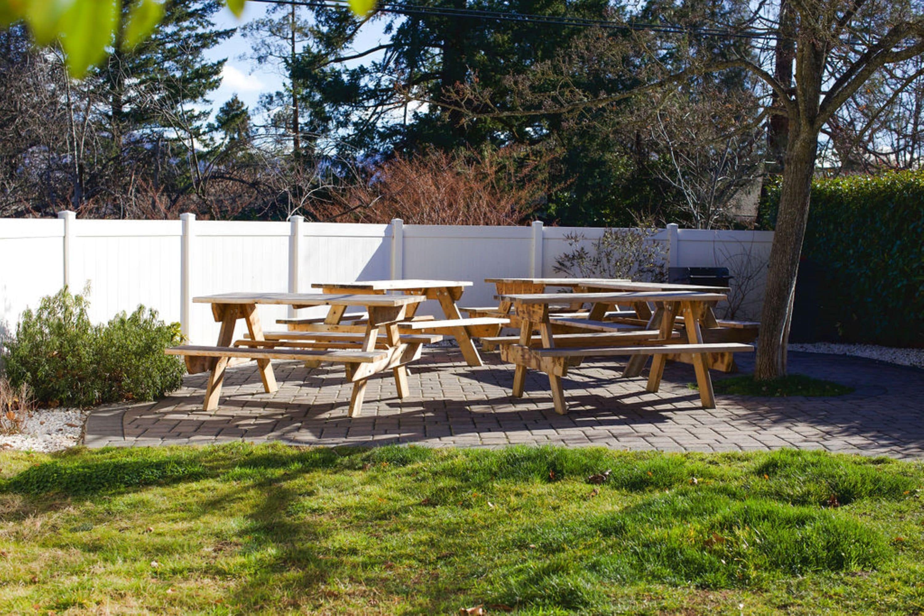 Un área de patio al aire libre con varias mesas de picnic dispuestas sobre una superficie pavimentada, rodeada por una cerca blanca y follaje.