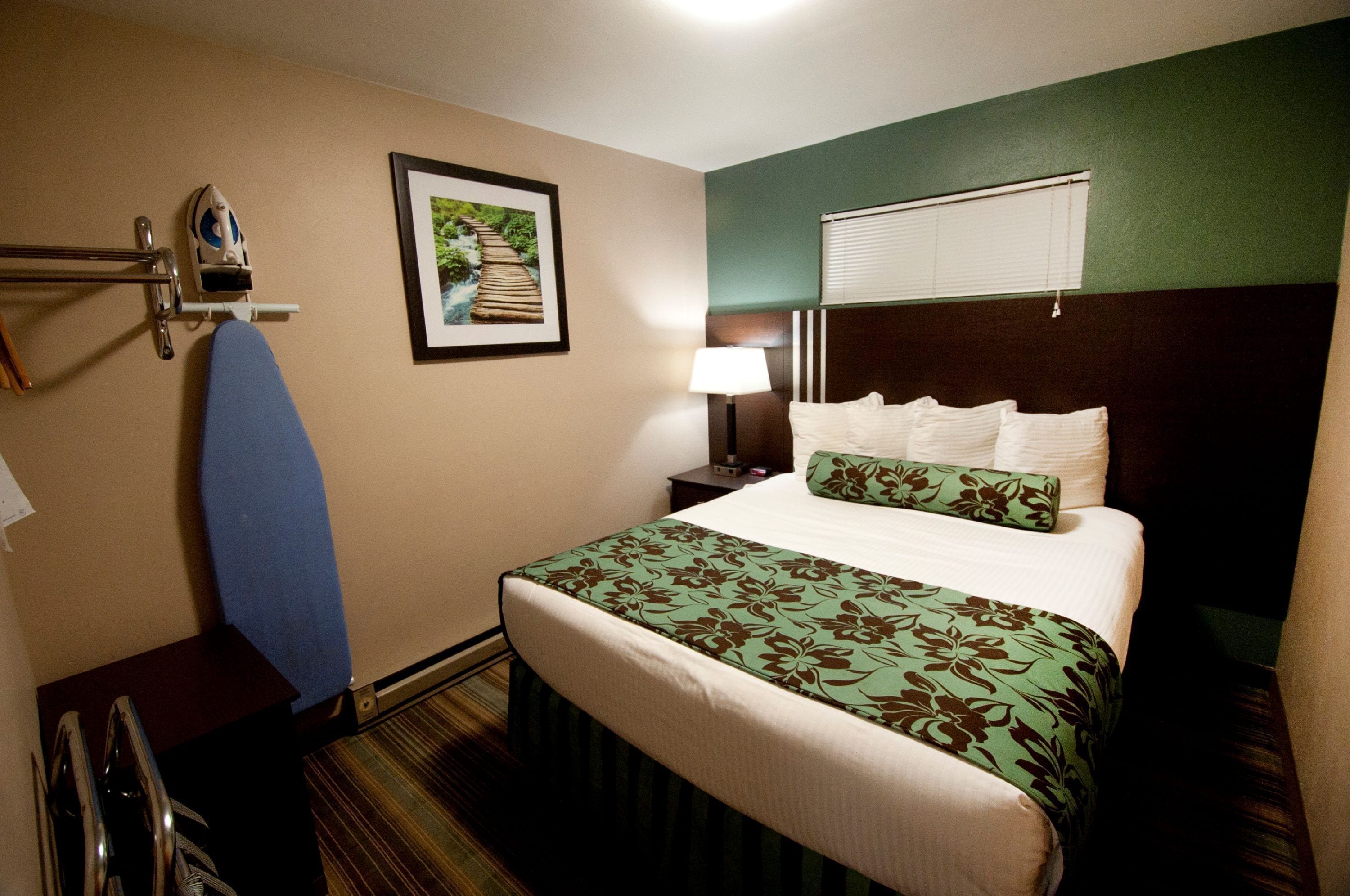Habitación de hotel con cama bien hecha, mesita de noche con lámpara, un cuadro en la pared y ventana con persianas.