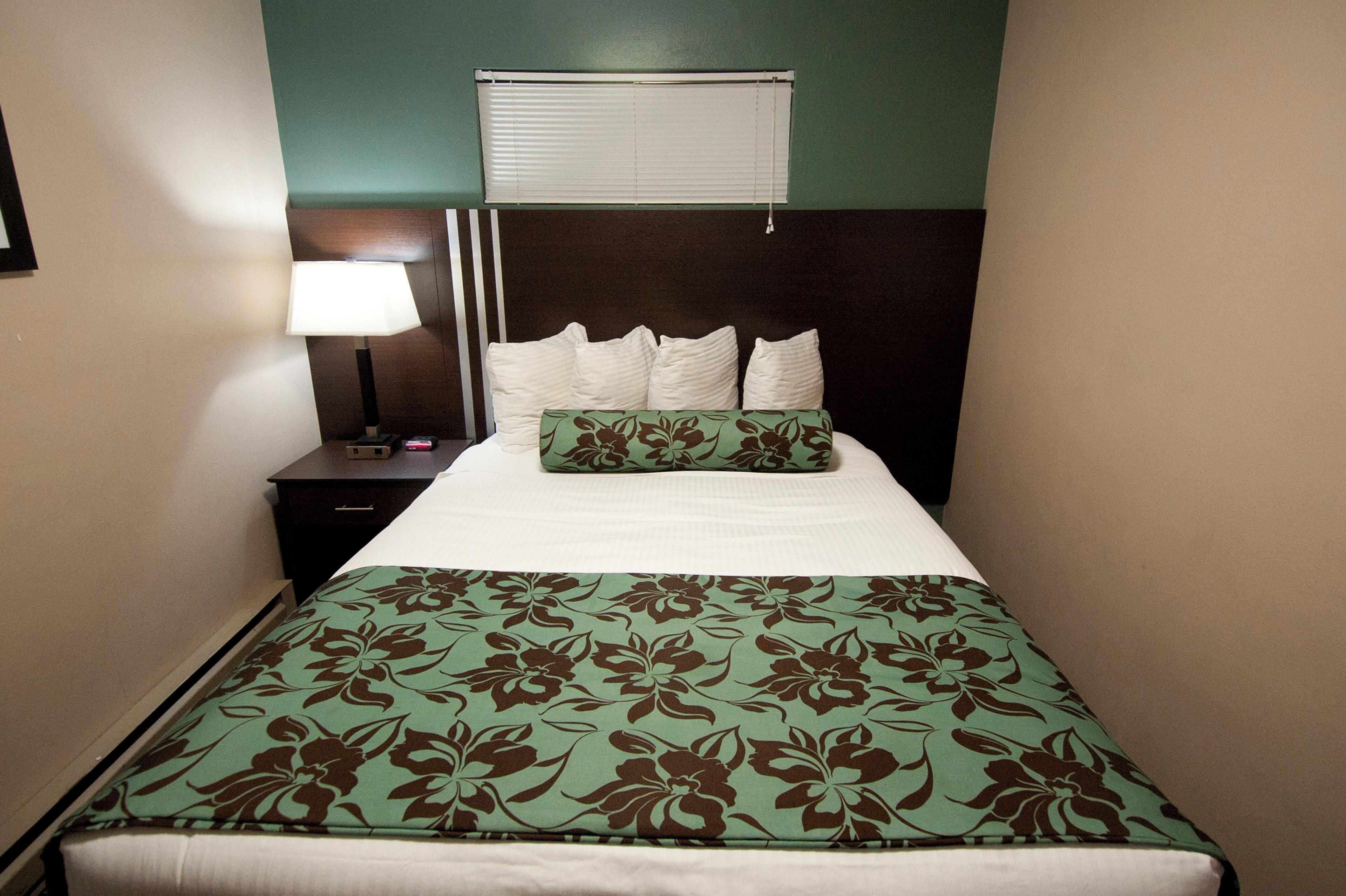 A neatly made bed with a floral bedspread and pillows, a bedside lamp, and a window with blinds.