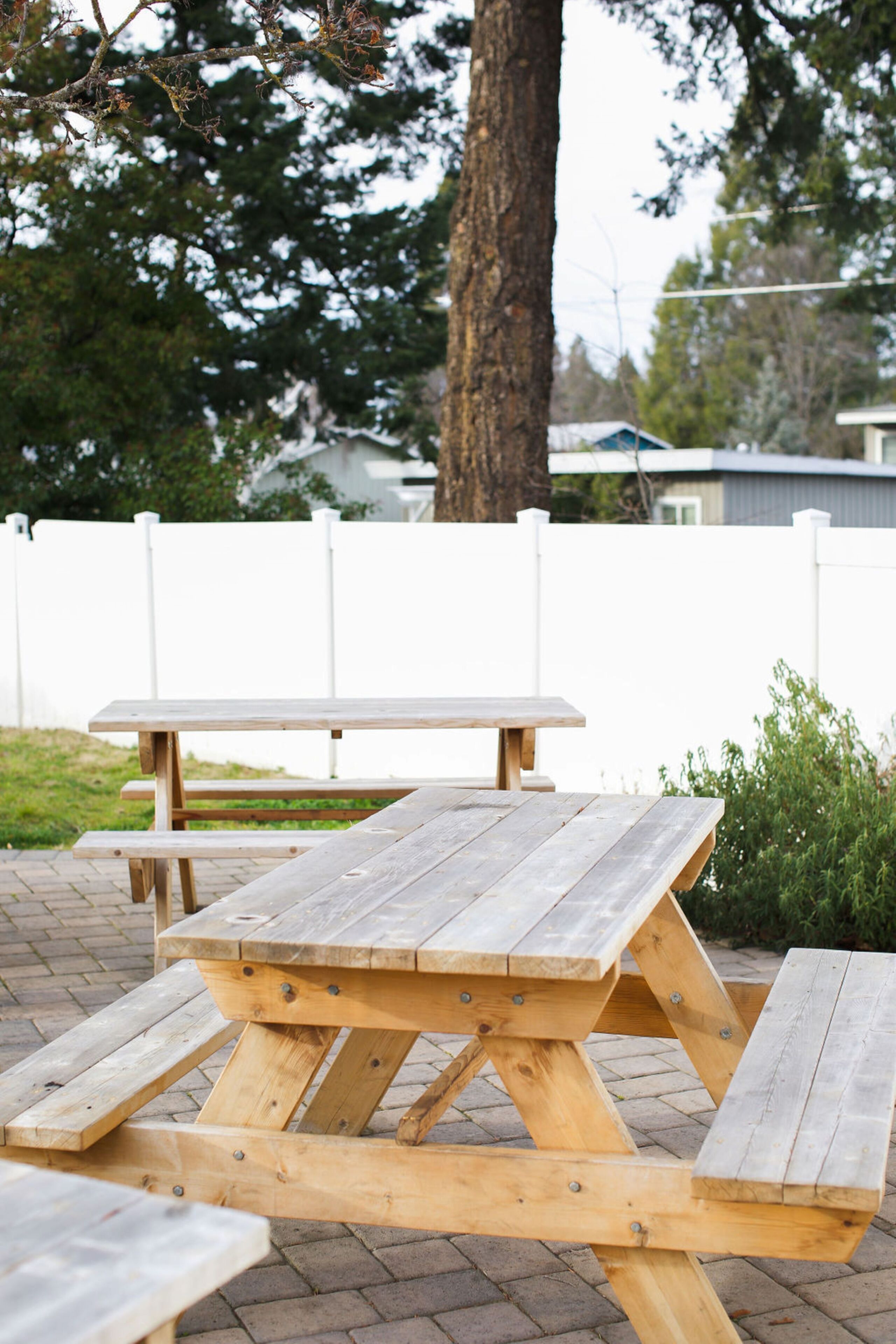 Une table de pique-nique en bois, avec des bancs, installée sur une surface pavée, avec une clôture blanche et des arbres en arrière-plan.