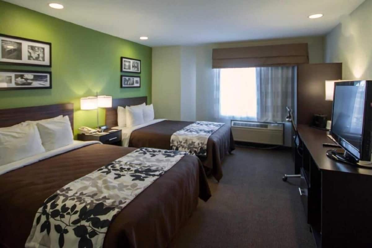 A hotel room with two beds, a desk with a television, an air conditioning unit, and framed pictures on the wall.
