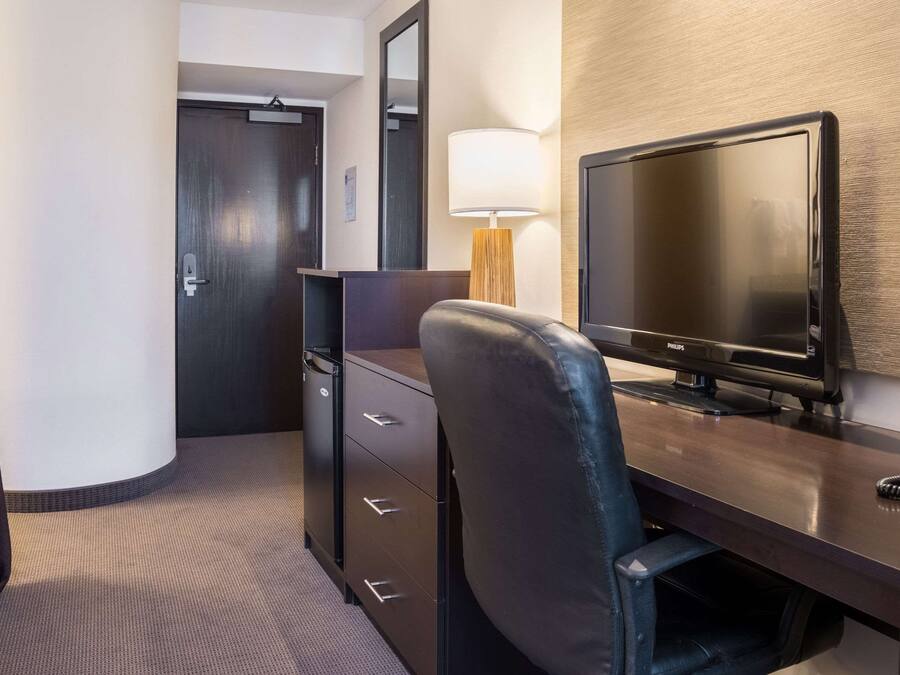 A hotel room with a desk, a television, a lamp, a wardrobe, and a door.