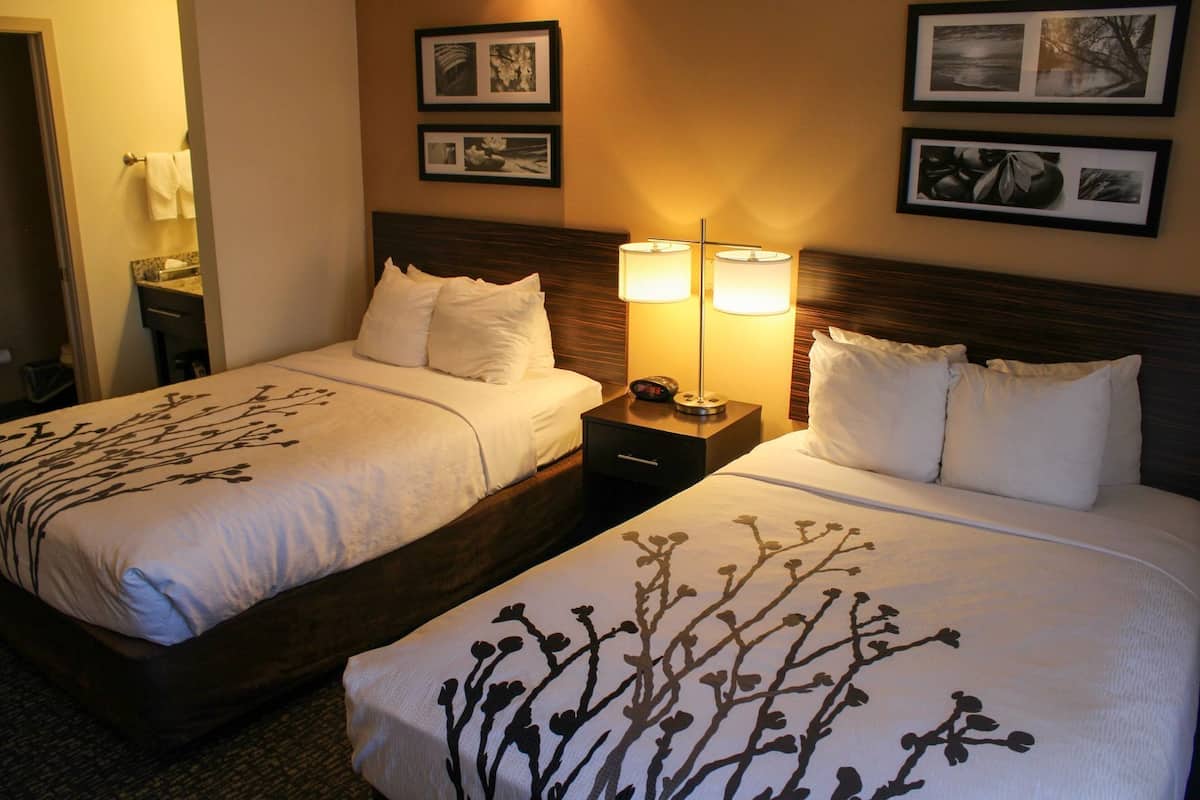 Duas camas de casal com roupa de cama branca e padrões florais pretos, luminárias de cabeceira e quadros na parede.