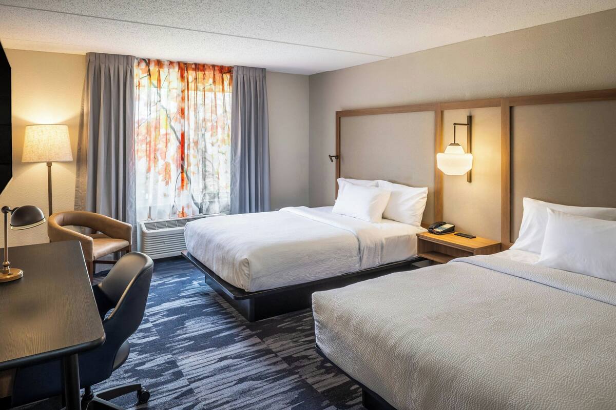 A hotel room with two beds, a desk, a chair, a lamp, and a window with curtains.