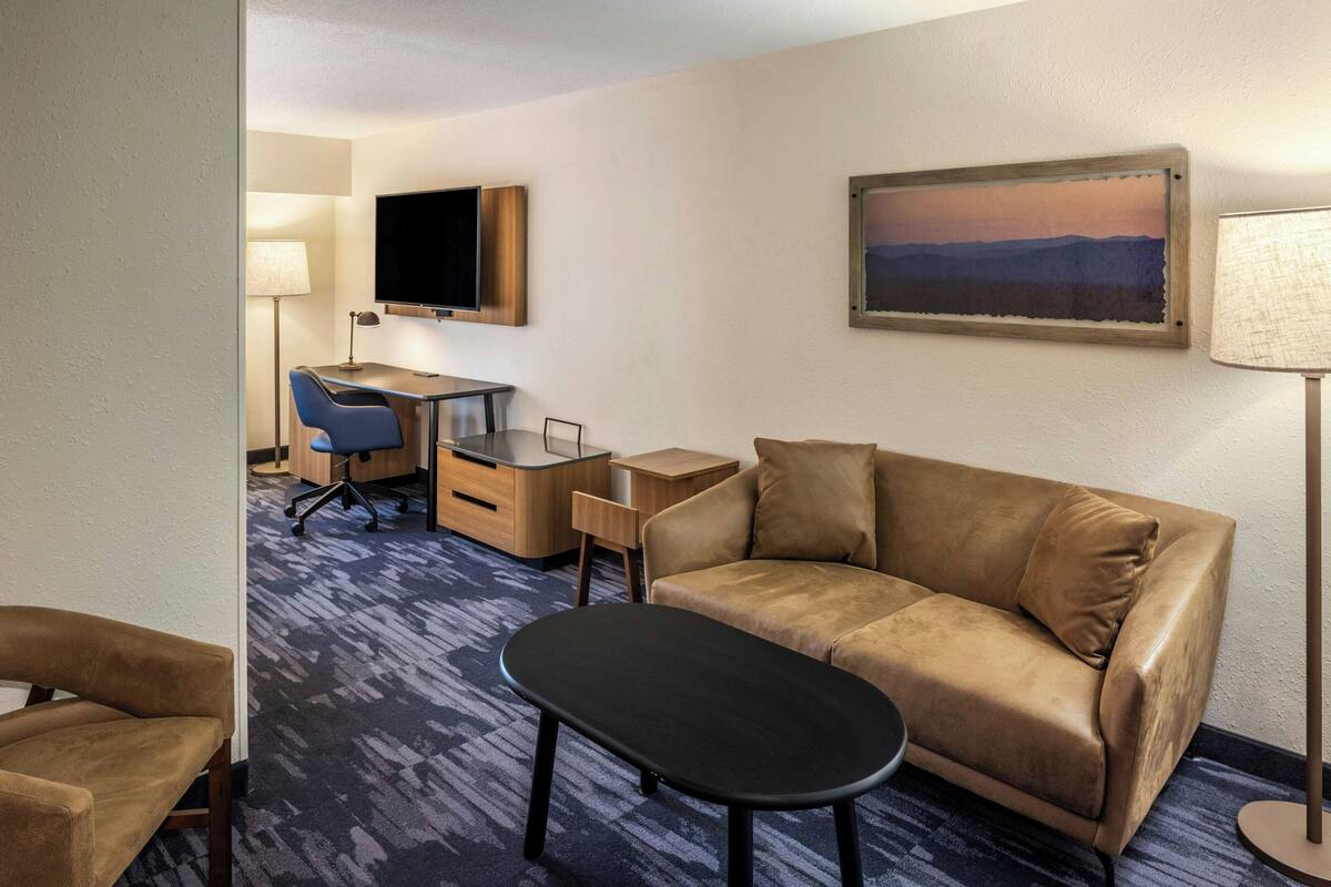 A hotel room with a sofa, a desk with a chair, a TV mounted on the wall, and a painting above the sofa.