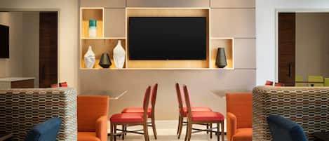 Un interior moderno con un televisor de pantalla plana montado en la pared, rodeado de estanterías con objetos decorativos. Hay dos sillas de madera con cojines rojos y un sofá estampado.
