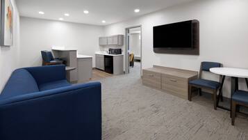 A modern hotel room with a blue sofa, a dining table with chairs, a kitchen area, and a flat-screen TV mounted on the wall.