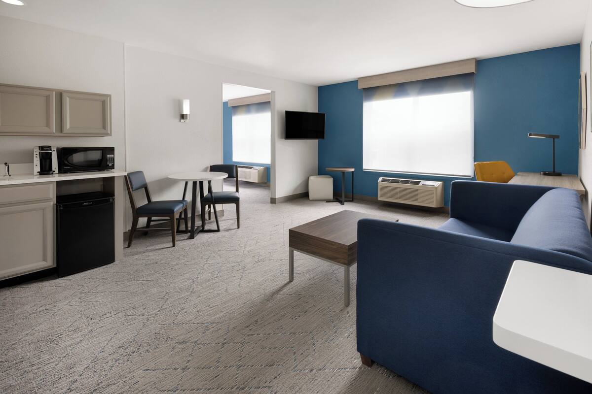 A modern hotel room with a kitchenette, a sofa, a dining area, and a TV.