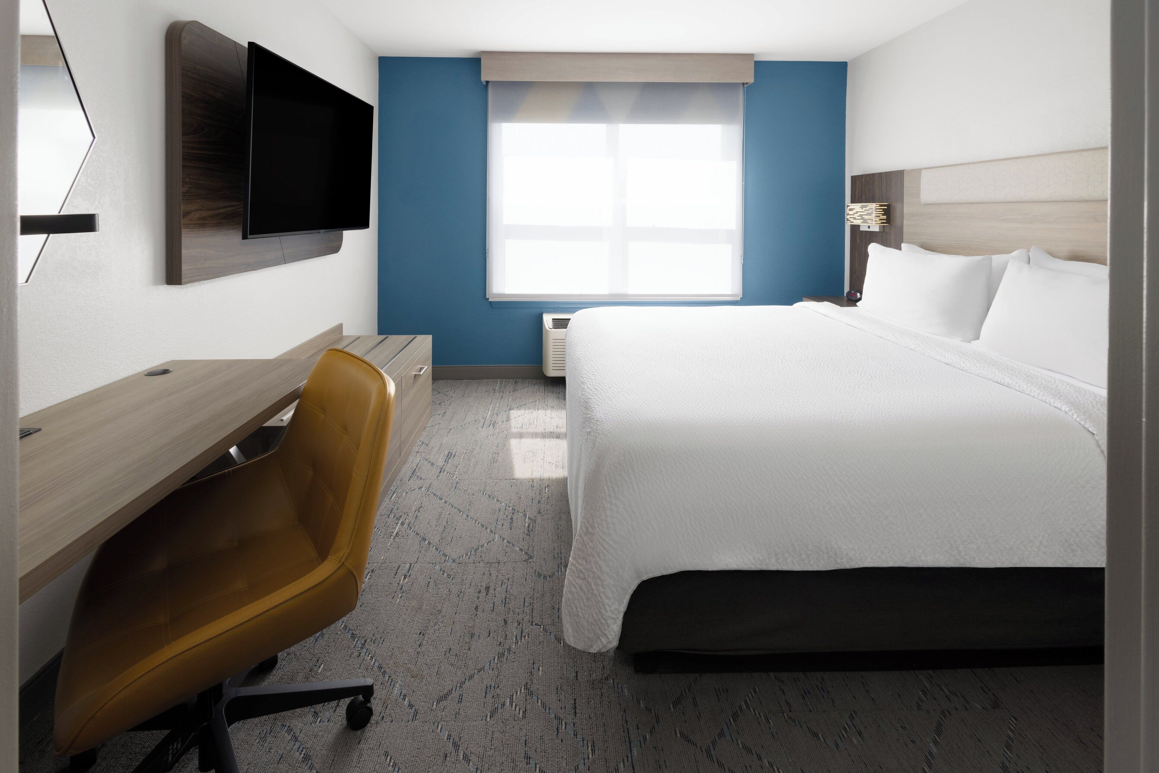 A hotel room with a bed, a desk, a chair, a TV mounted on the wall, and a window with blinds.