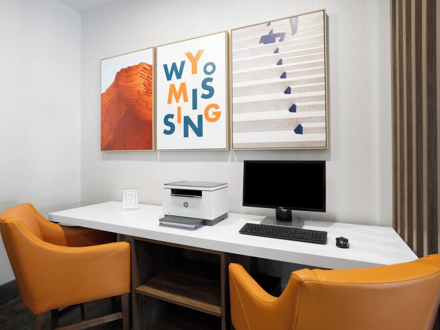 Een moderne kantoorruimte met een wit bureau, twee oranje stoelen, een printer, een monitor en drie ingelijste kunstwerken aan de muur.