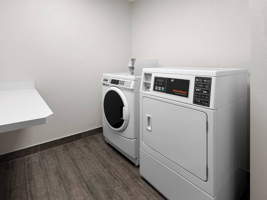 A laundry area with two front-loading washing machines and a drying machine, a small white shelf, and a dark wooden floor.