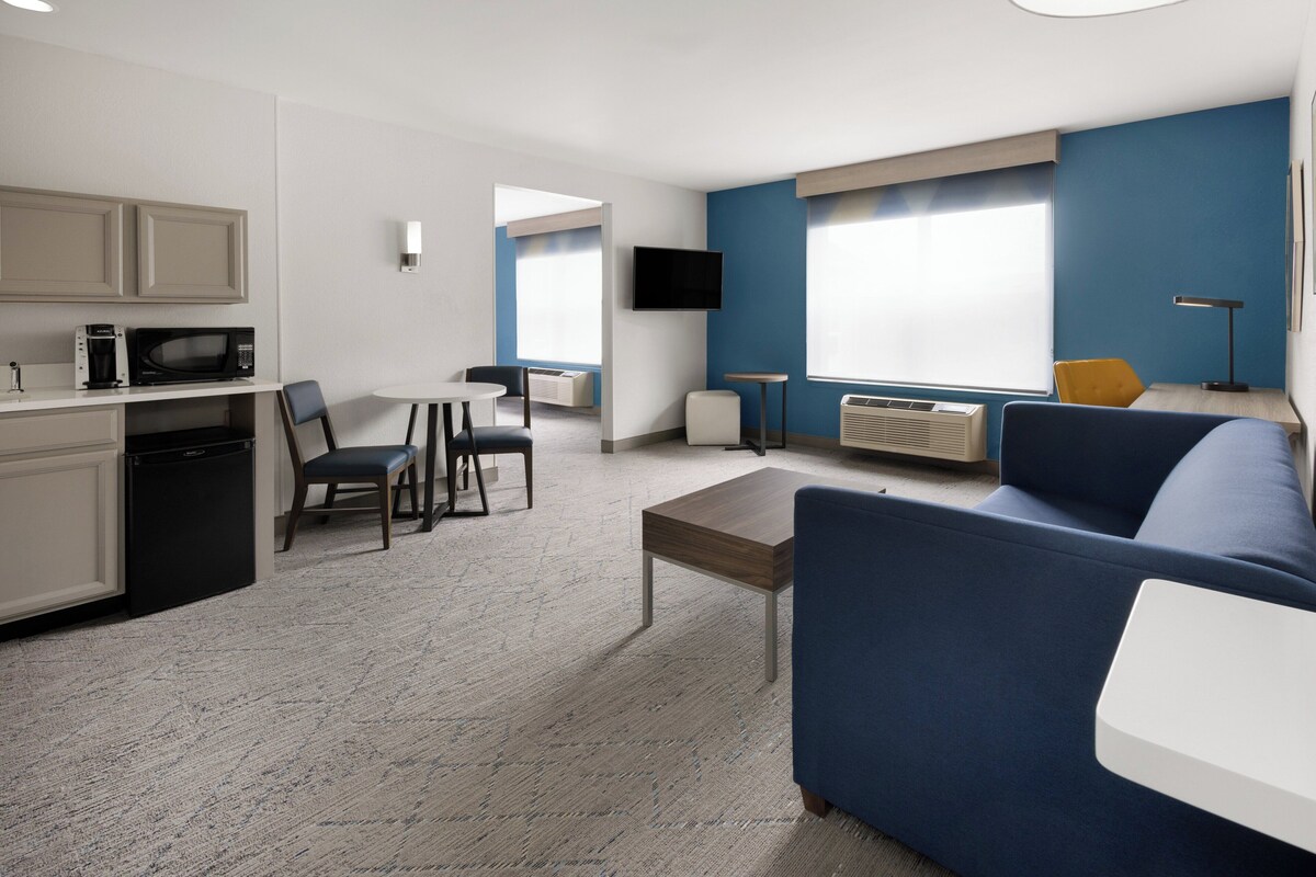 A modern hotel room with a kitchenette, a sofa, a dining area, and a TV.