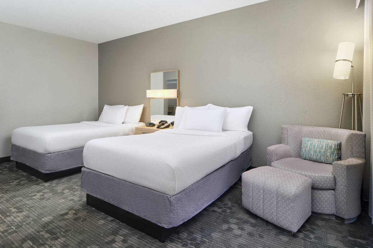 A hotel room with two beds, a chair, a lamp, and a mirror.