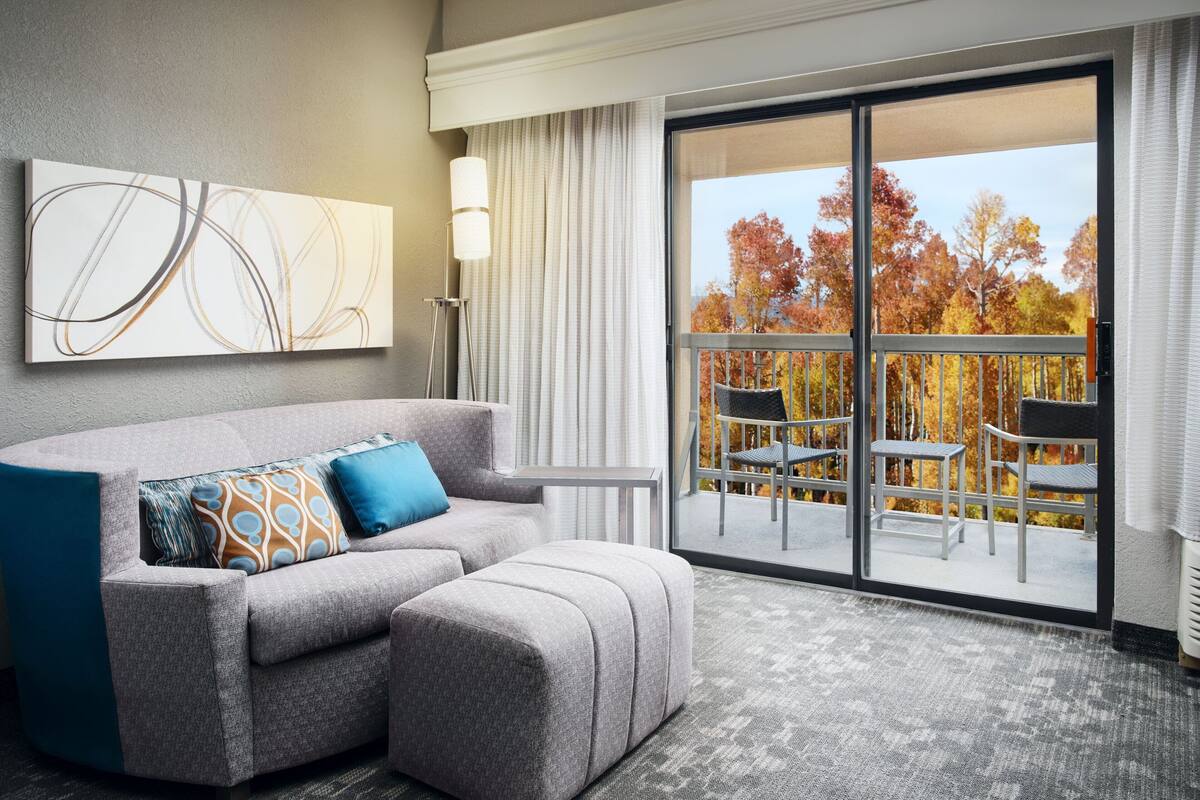 A modern hotel room with a grey sofa, ottoman, and a large abstract painting on the wall. A floor lamp is visible, and the room has a large sliding glass door leading to an outdoor balcony with chairs.