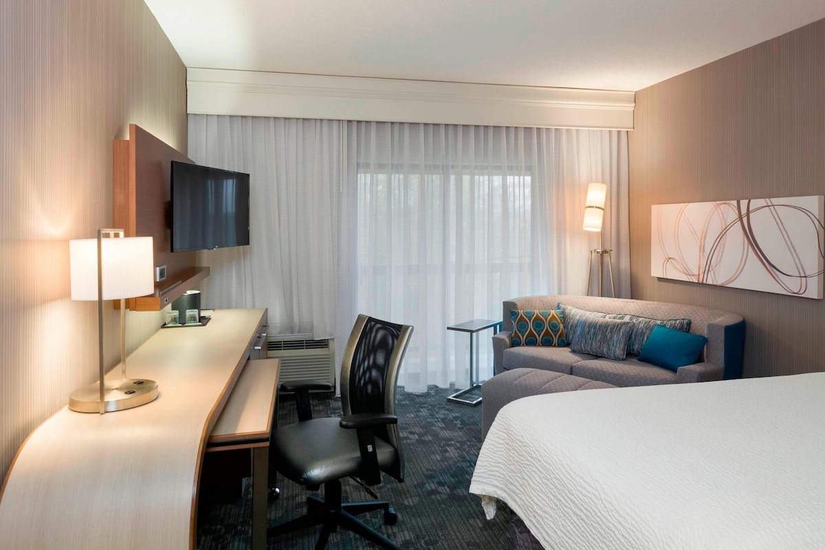 A hotel room with a bed, a desk with a chair, a television, a small table with a chair, and a window with curtains.