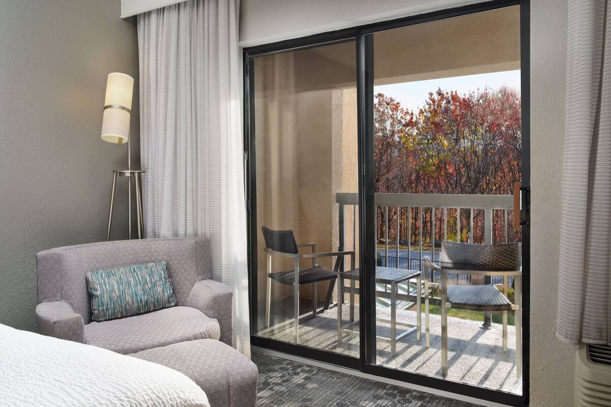 A hotel room with a large sliding glass door leading to an outdoor patio with a view of autumn trees.