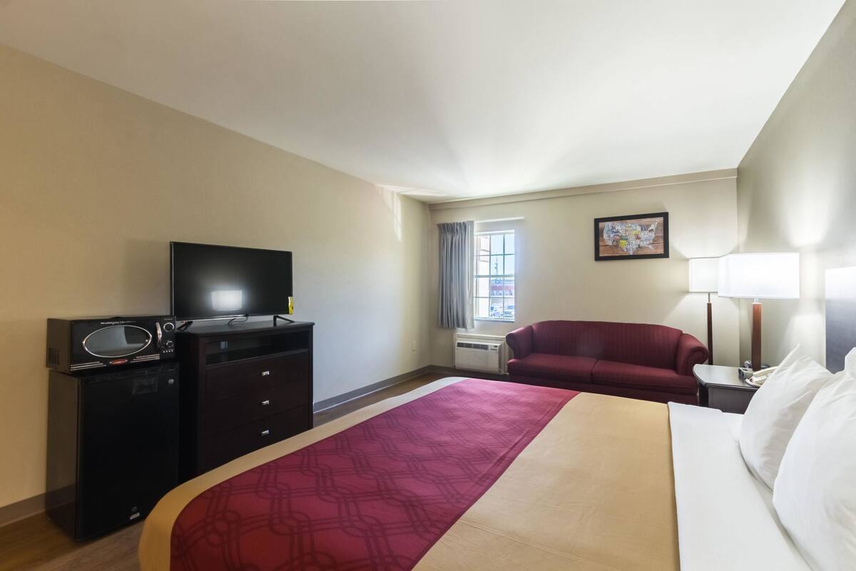 A hotel room with a bed, a TV, a desk with a lamp, a red couch, and a window with curtains.