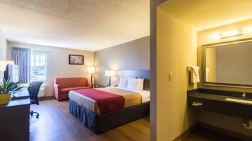 A hotel room with a bed, a desk, a chair, a plant, and a bathroom with a sink and mirror.