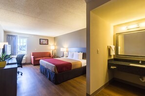 A hotel room with a bed, a desk, a chair, a plant, and a bathroom with a sink and mirror.