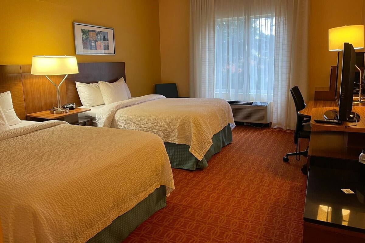 A hotel room with two beds, a desk with a computer, a chair, a lamp, and a window with curtains.
