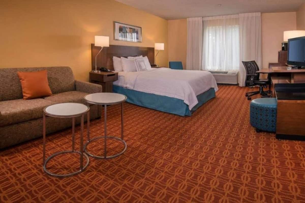 A hotel room with a bed, a sofa, two small tables, a desk with a chair, and a television.