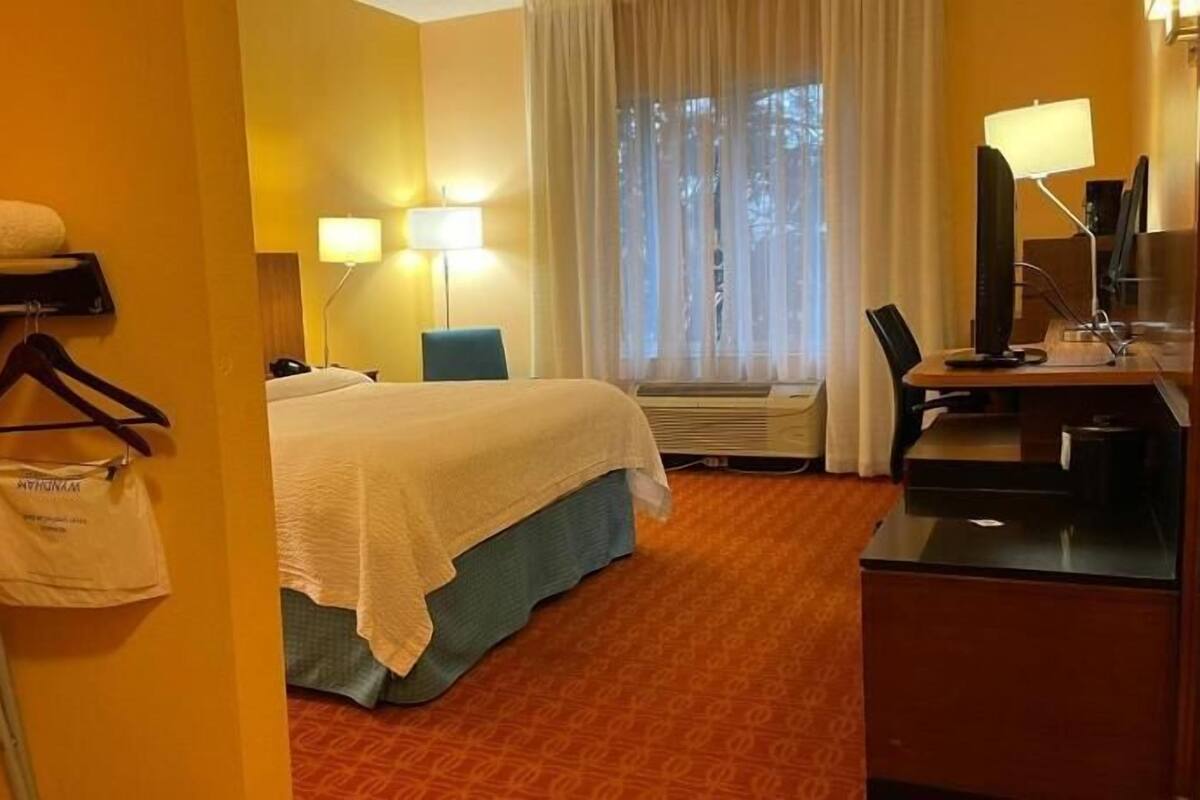 A hotel room with a bed, a desk with a lamp, a chair, a desk lamp, a wall-mounted heater, and a window with curtains.