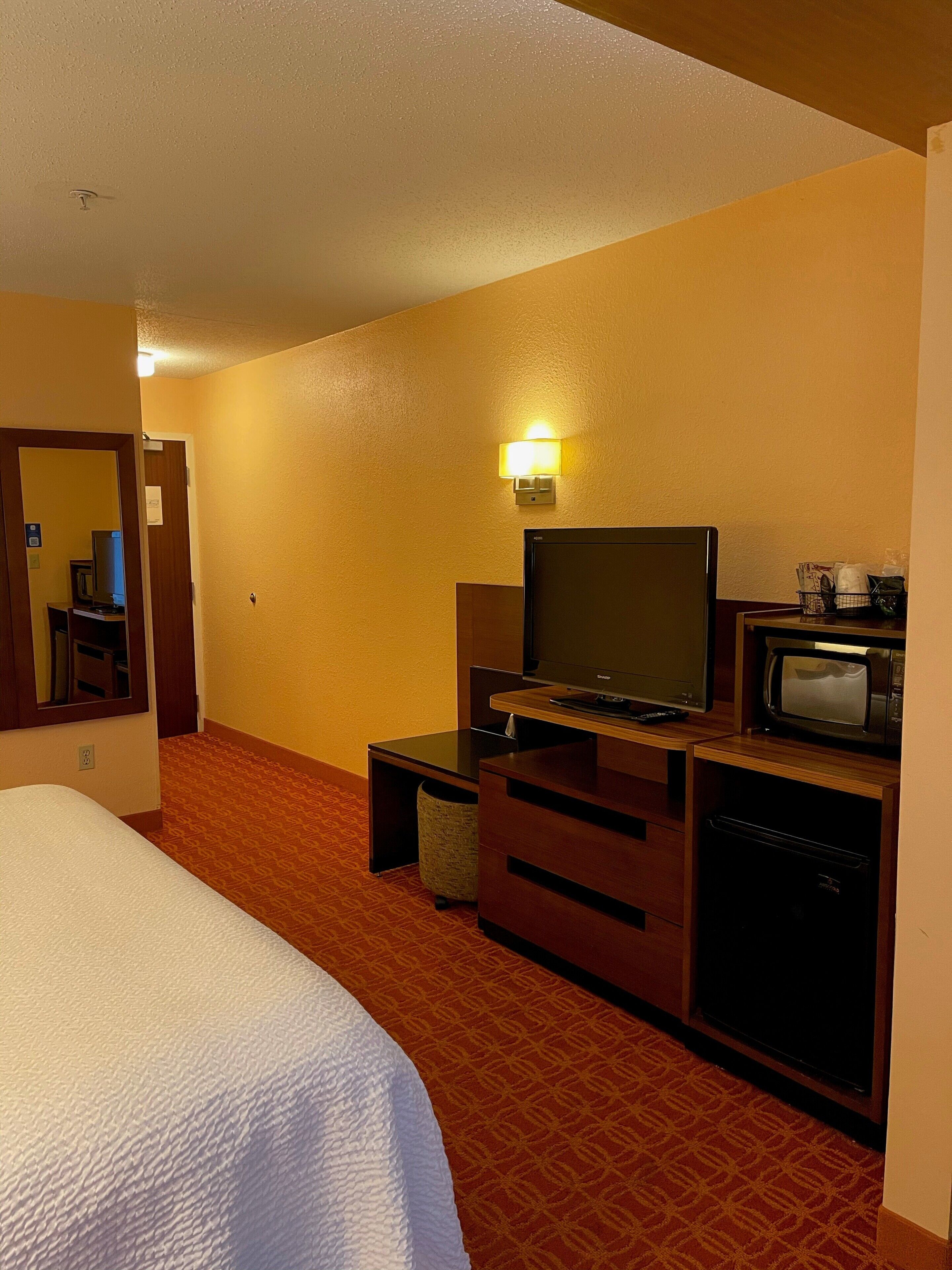 A hotel room with a bed, a television, a microwave, a small table, and a wall-mounted lamp.