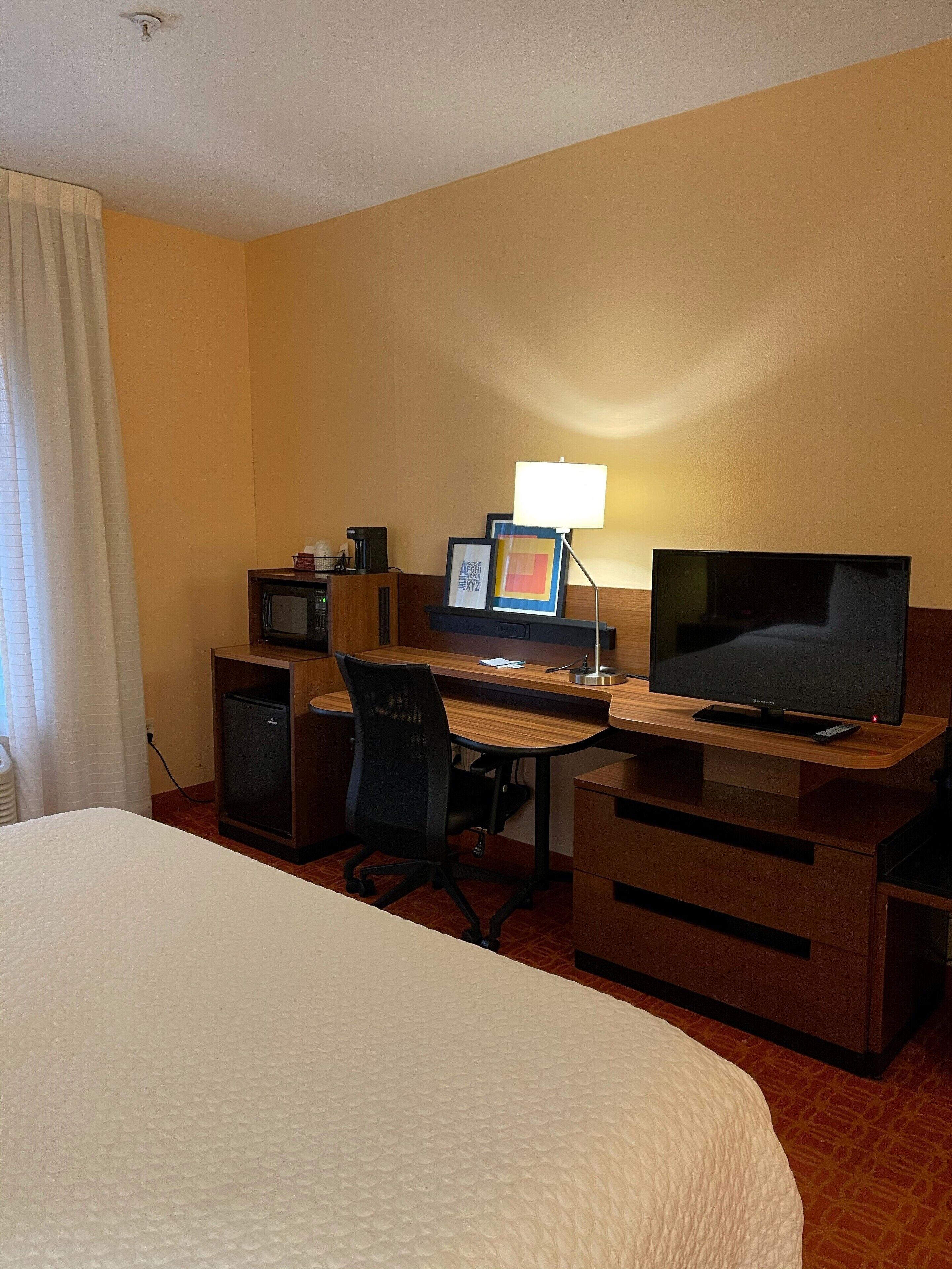 Una habitación de hotel con una cama, un escritorio con computadora, televisor y un refrigerador pequeño.