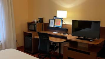 Una habitación de hotel con una cama, un escritorio con computadora, televisor y un refrigerador pequeño.