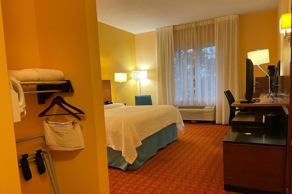 A hotel room with a bed, a desk with a computer, a chair, a desk lamp, a window with curtains, and a wall-mounted heater.