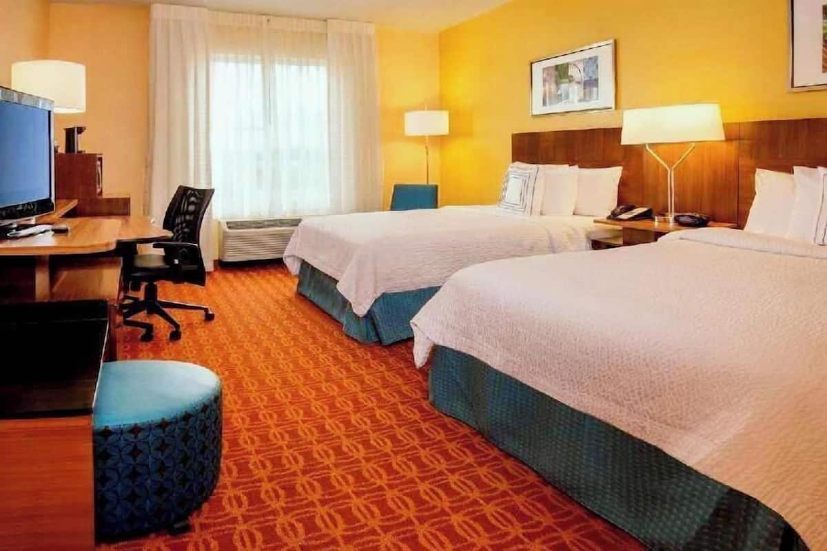 A hotel room with two beds, a desk with a computer, a chair, a lamp, and a window with curtains.