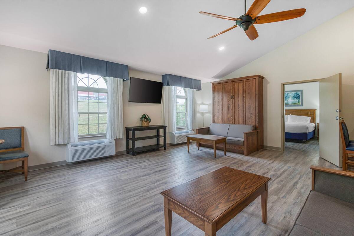 Un vaste espace de vie doté d’un ventilateur de plafond, d’une grande fenêtre avec des rideaux, d’une table en bois, d’un canapé et d’un téléviseur fixé au mur.