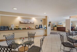Un vestíbulo de hotel con mostrador de recepción, un área de comedor con sillas de metal y una mesa de vidrio, y una sala de estar con sofá y sillones.