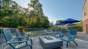 Een terras buiten met Adirondack stoelen, een vuurkorf en blauwe parasols.