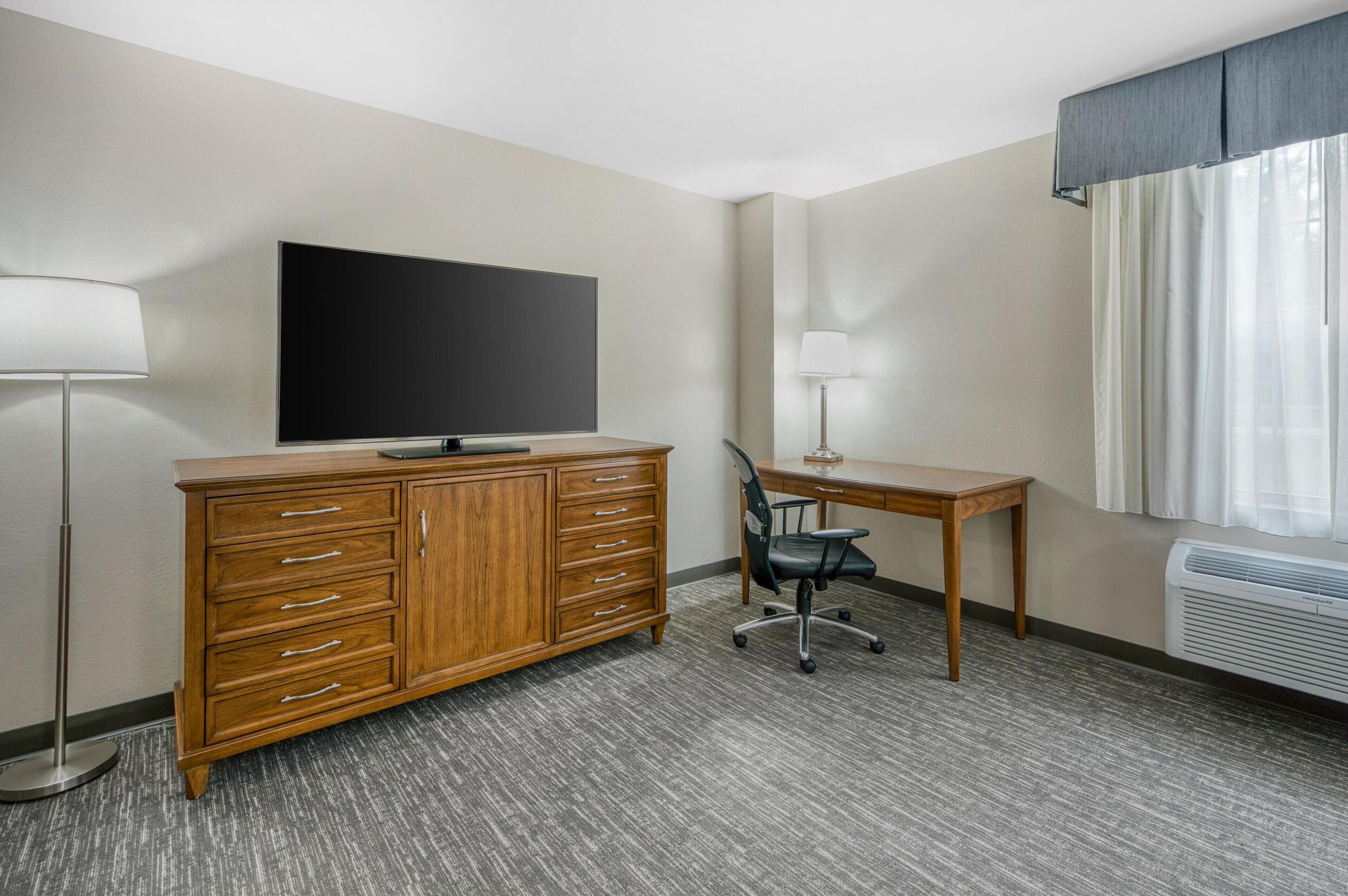 Hotelkamer met een houten dressoir, een flatscreen televisie, een bureau met stoel, een staande lamp en een raam met gordijnen.