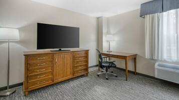 Hotelkamer met een houten dressoir, een flatscreen televisie, een bureau met stoel, een staande lamp en een raam met gordijnen.